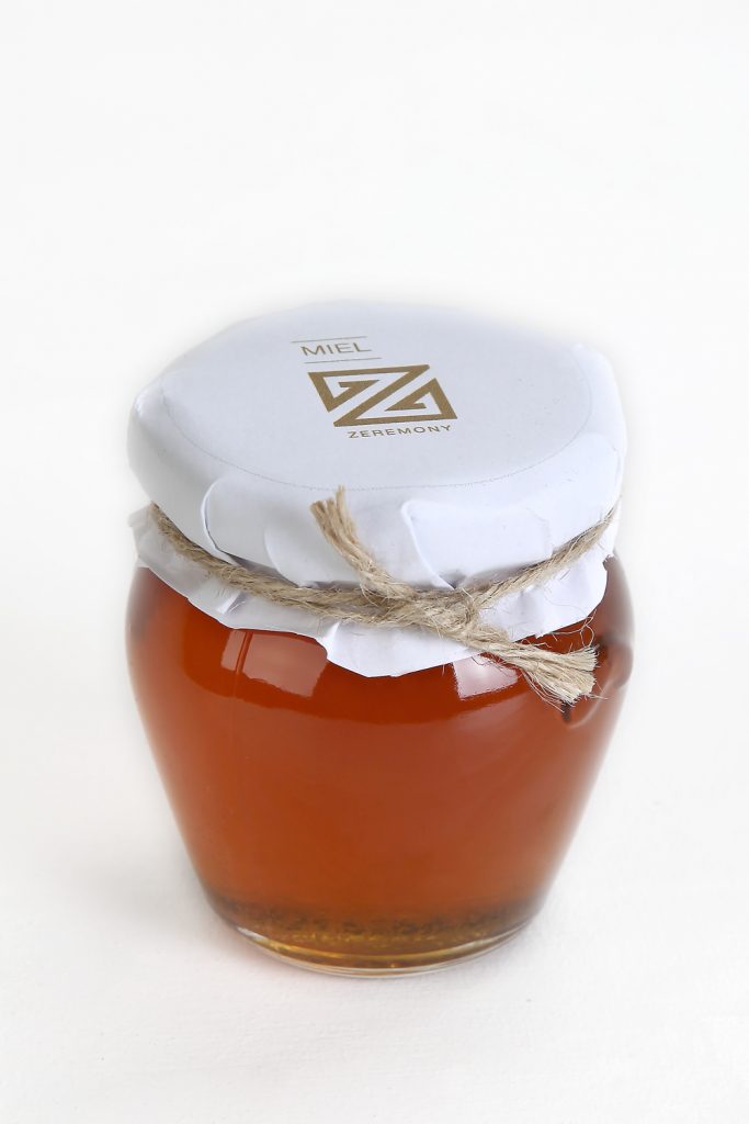 Esta miniatura de miel tiene forma ovoide achatada con base redonda. Está decorado con todo el capricho con un papel en el que se puede ver la elegancia de la marca Zeremony y un lacito.